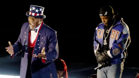 Getty Images Samuel L Jackson as Uncle Sam during Kendrick Lamar's halftime show. Jackson wears blue tails with stars on the lapels over a red waistcoat, white shirt and black bow tie. His outfit is completed by a blue top hat decorated with the stars and stripes of the US flag. He holds his hands out, mid speech, while Kendrick stands behind him, looking down with his bands folded in front of him.