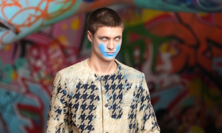 Model with short-cropped brown hair, white contact lenses and a blue painted U-shape running from cheekbone to cheekbone around their jaw