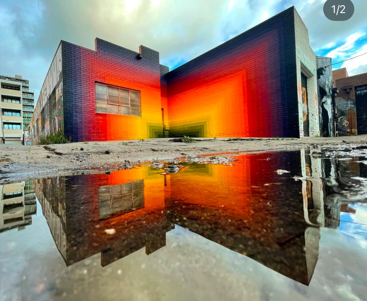 building painted with red and orange abstract mural reflected in water on ground. Street art