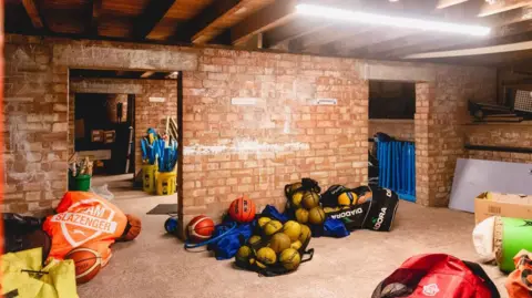 Broadleaf Partnership Trust A sports storage room with brick walls and bags of balls and other sporting equipment piled up on the floor.