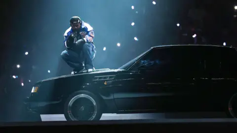 Getty Images Kendrick Lamar during his halftime show at the Super Bowl. The stadium is dark with the rapper lit by a single spot light as he crouches on the bonnet of a black muscle car.