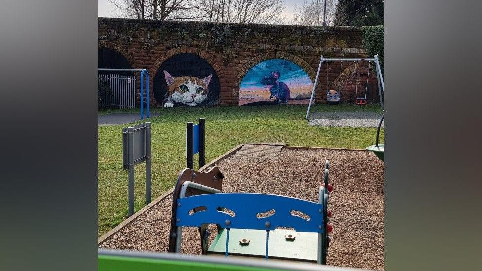 The two murals in the brick arches are in the background. In the foreground is a play area with bark on the floor and a pair of swings. Other equipment is blue and green.