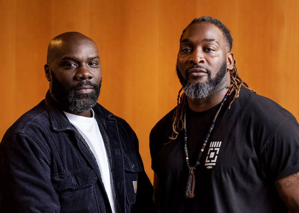 Two Black men are pictured from the waist up, gazing evenly into the camera. Both have trimmed beards and wear black street clothes. Asante, on the left, is bald-headed; Sandy has long thin dreadlocks.