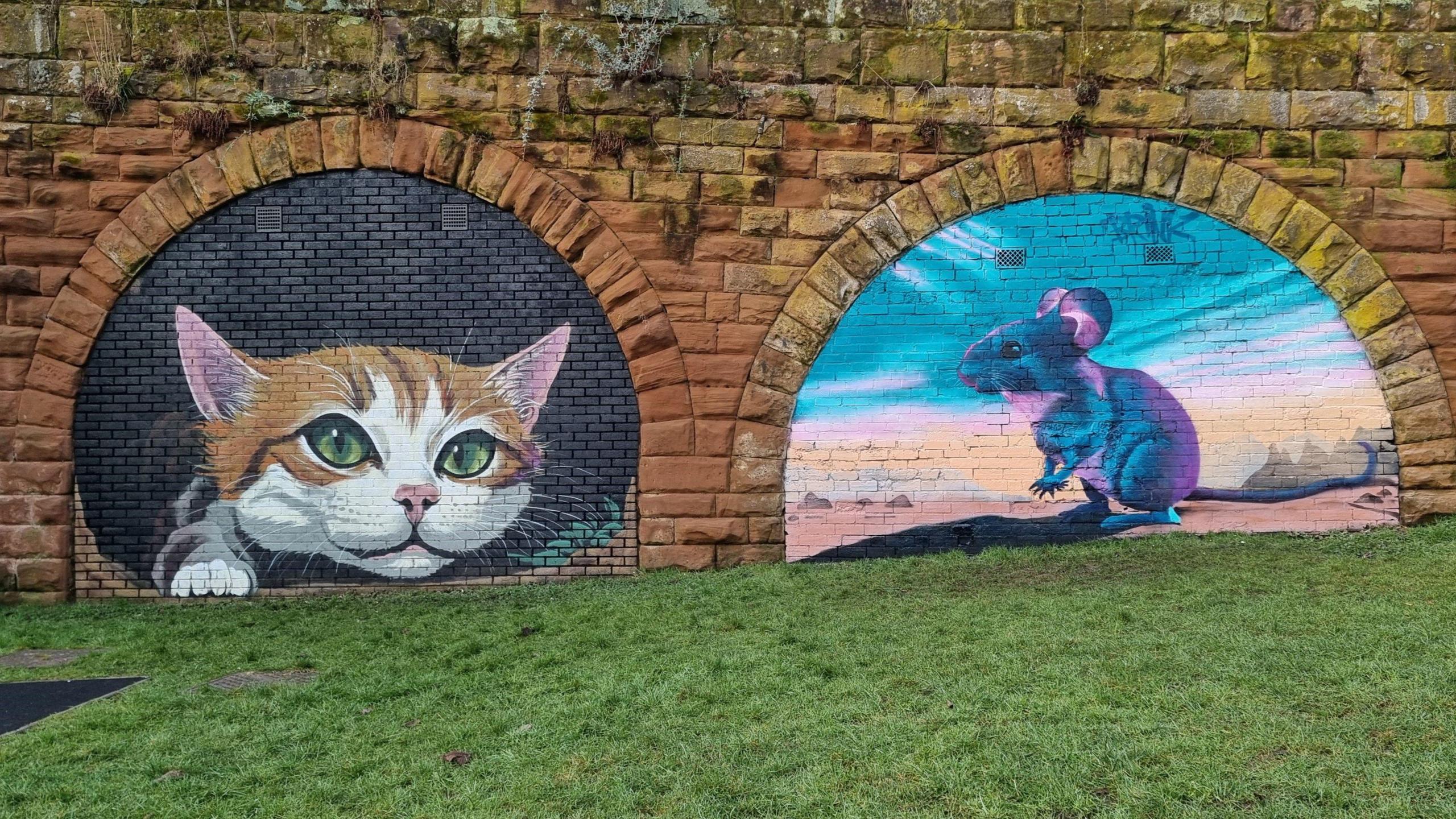 Two filled in brown brick archways that have been painted on. The mural on the left is a ginger and white cat that appears to be climbing out of the bricks. It has green eyes and there are some leaves painted next to it. On the right is a painting of a grey mouse, with light shining on it. The mouse is standing in a desert-like landscape with hills and rocks in the background.