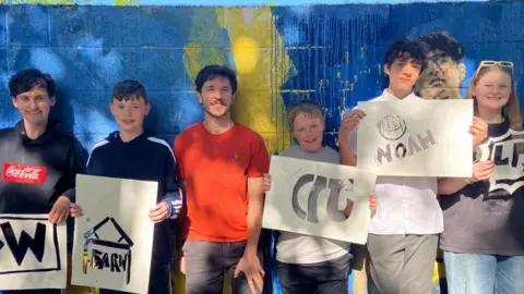 Fiona Callow/BBC A group of children hold up pieces of paper with cut-outs of their graffiti tags. They are standing in front of a brick wall spray-painted blue and yellow.