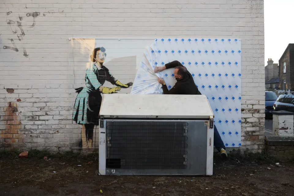 A worker covering a Banksy artwork depicting a woman in a 1950s-style dress with a protective screen.