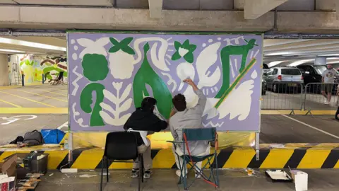 BBC Two people sat down on chairs painting a canvas inside a car park - it has green white and purple coloured designs on it.