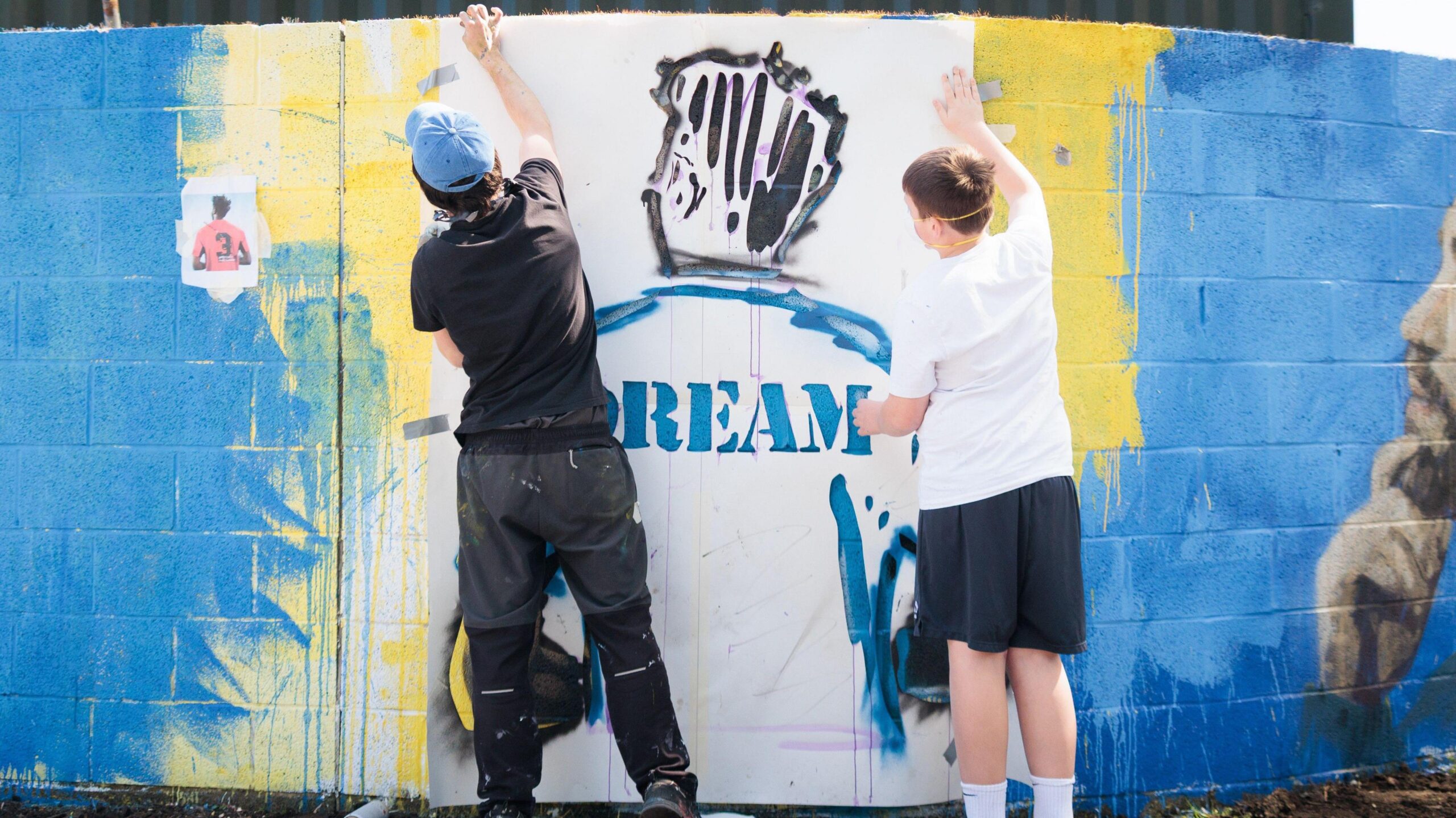 Two figures work on a blue spray-painted brick wall, filling in the outline of the back of a football player's shirt.