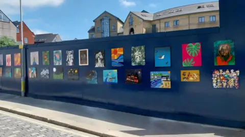 Mike Vosper Multiple bright artworks on dark blue hoardings, the sky in the background is blue.