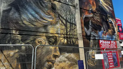 Alice Toon/ BBC A mural on the side of a concrete wall, on the left a design of the right side of a statue's face coloured in yellow and grey, on the right an image of an eagle with wings spread holding an oil lamp. There is a phone repair advertisement in the bottom right corner and scaffolding in front of the mural.