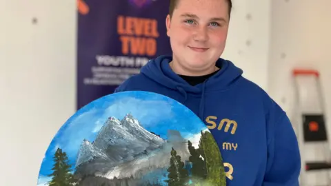 Jon Wright/BBC A young person in a blue hoody holds a circular canvas with a painting of mountains, blue sky and trees,