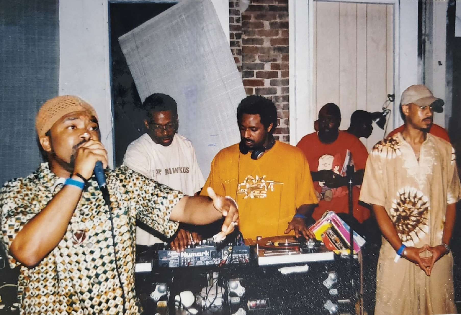 Gene Brown raps onstage while standing in front of a group of about five guys dressed in 1990s hip-hop gear.