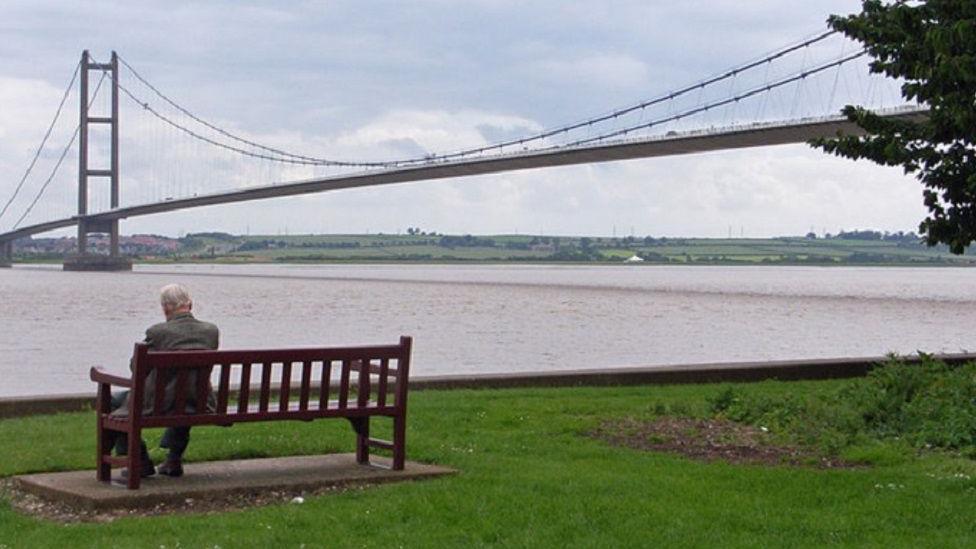 Humber Bridge viewing area on Hessle Foreshore, with a man sat on a bench on a grassy area next to the river