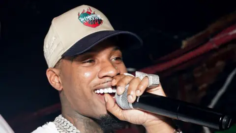 Getty Images A man performs on stage. He's smiling as he cups his hand around a microphone held close to his mouth. He's wearing, large, chunky rings encrusted with jewels and a wide silver chain around his neck. He wears a sandy coloured baseball cap with a navy peak and the word