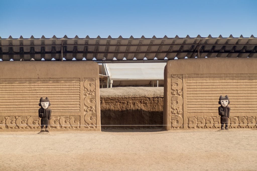 Adobe walls with carved figures and reliefs at Chan Chan archaeological site under protective roofing.