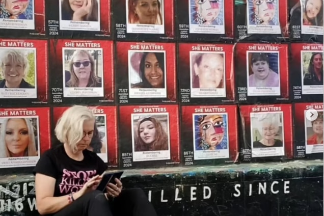 A woman with chin length blonde hair sits on the ground looking at her phone against dozens of photos of woman on a wall.