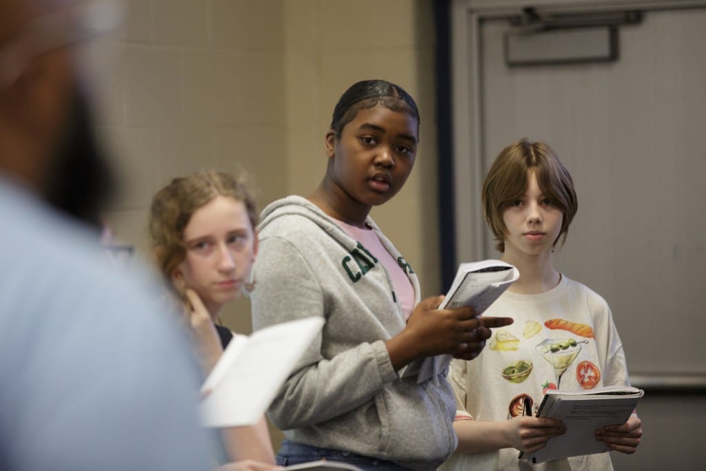 Three middle school girls stand with notebooks in hand listening to Trevon Hobson's feedback.