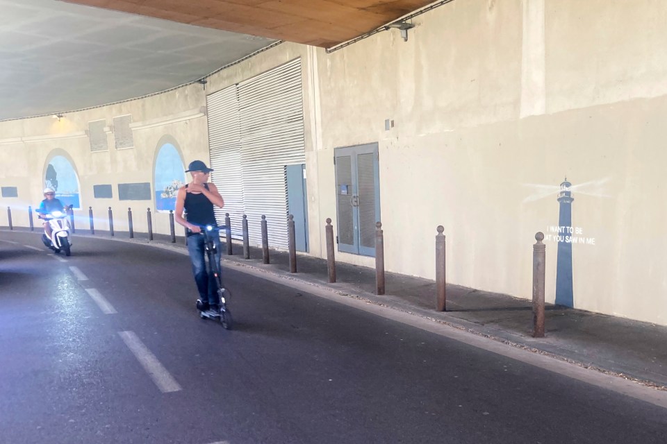 Two people riding scooters under a bridge with a mural.