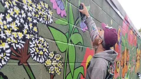 Nick Ballard A street artist wearing a purple baseball cap and grey hoodie top spray paints a flower onto a brick wall.