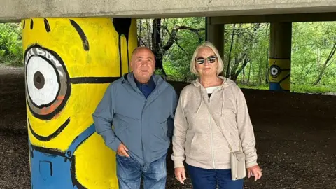 Pete Storozuk and Julie Robinson standing by a motorway stanchions painted to look like a Minion - the yellow cartoon creations featured in the Despicable Me movies.