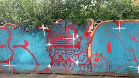 Chloe Aslett/BBC Temporary boards painted turquoise blue. Red lines create graffiti-style lettering but it is hard to tell what it says. In the centre is an alien-style creature - the red lines give its body a brain-like texture, but somehow its little face makes it quite cute. There are white stars dotted around the board.