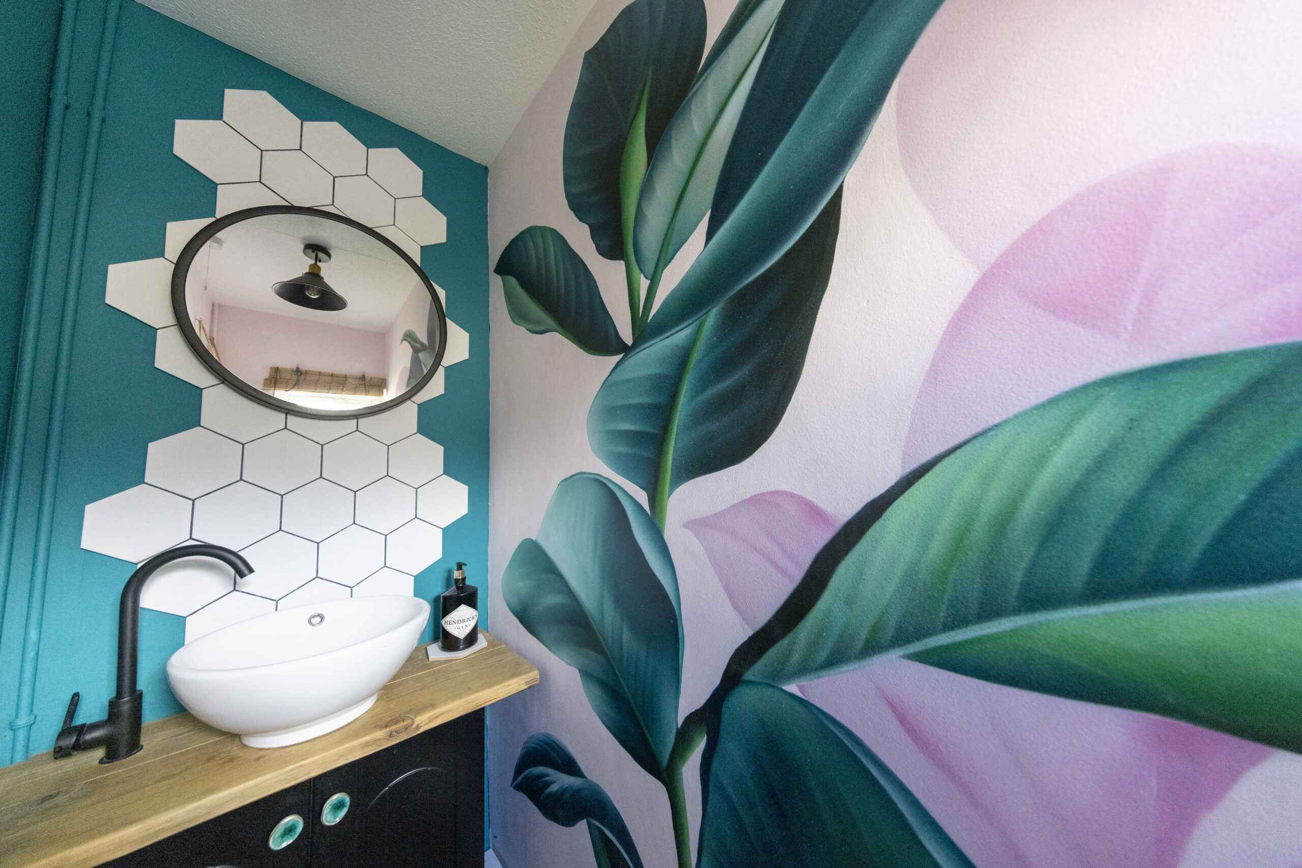 A painted bathroom with teal walls, hexagon tiles, and a large leaf mural.