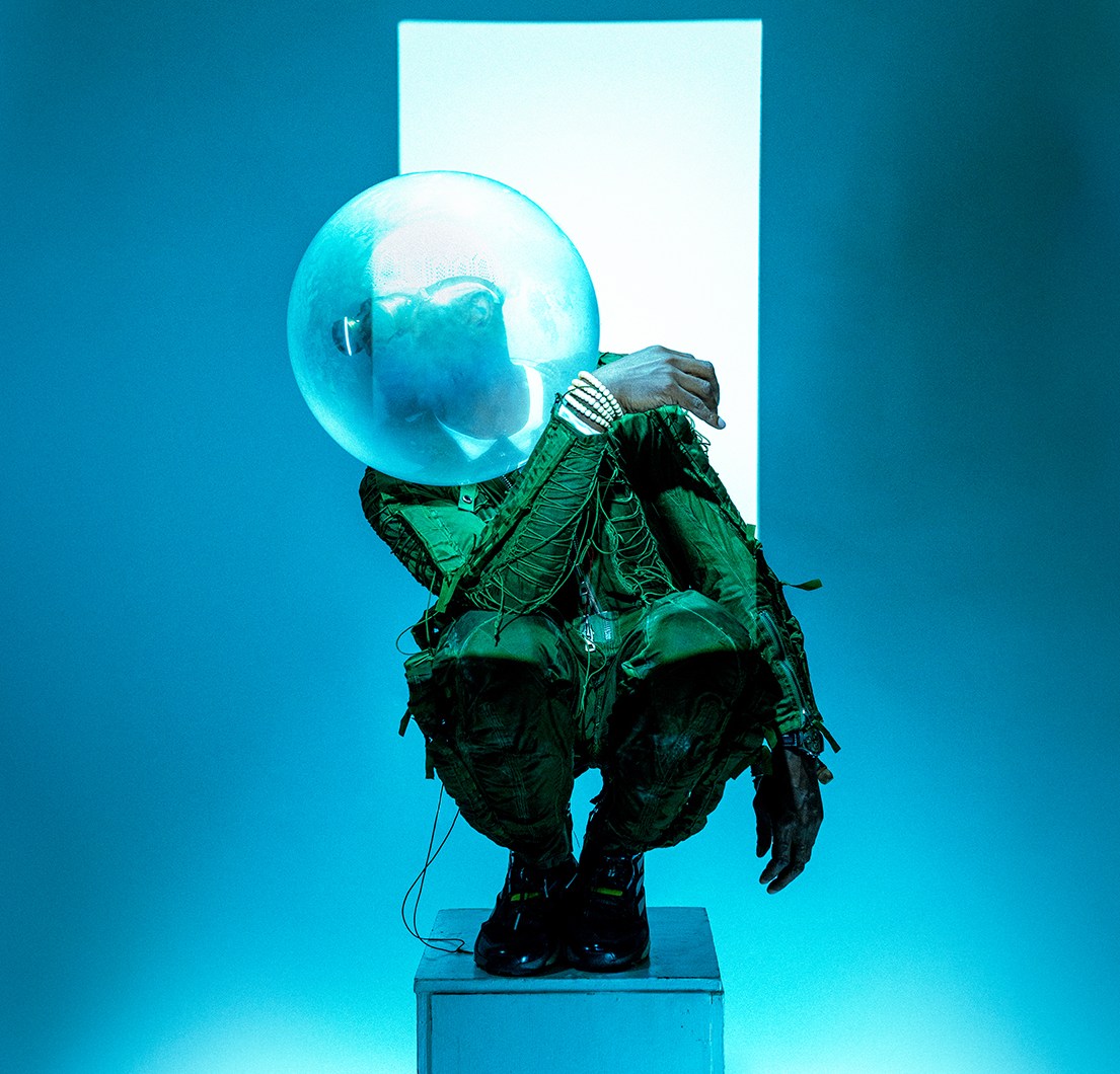 Person in green suit and bubble helmet crouching on a box.