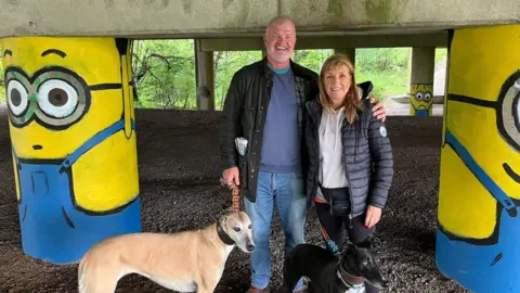 Eric and Lindsey Kerr holding the leads of their dogs as they stand in between two motorway stanchions painted to look like Minions - the yellow cartoon creations featured in the Despicable Me movies.