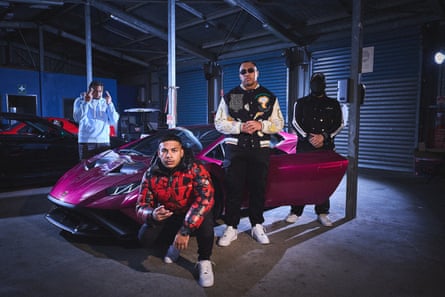 Four men, one in a full balaclava, stand around a purple sports car in a garage, striking poses