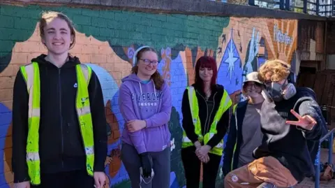 Lick of Paint/Instagram Five people in their late teens or early 20s. They are all smiling at the camera with their work behind them. It looks a sunny day.
