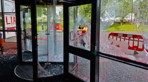 BBC Aftermath of an attack by protestors on a BBC building. A revolving door and panels with shattered glass, some of which has also fallen onto the floor, and red spray paint. Outside, barriers have been erected.
