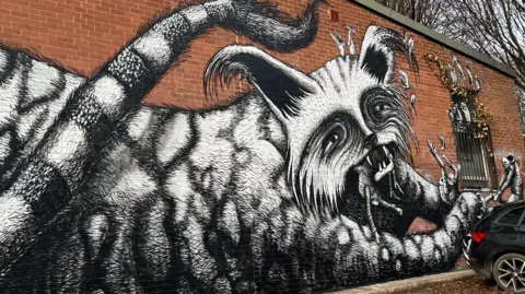 Xanthe Palmer/BBC The mural, which is on a brick wall. A black and white depiction of a giant, grotesque cat who is trying to eat a group of human beings. One of the people is hanging from its mouth. Another is being attacked by its claws, whilst a third tries to pull them free.