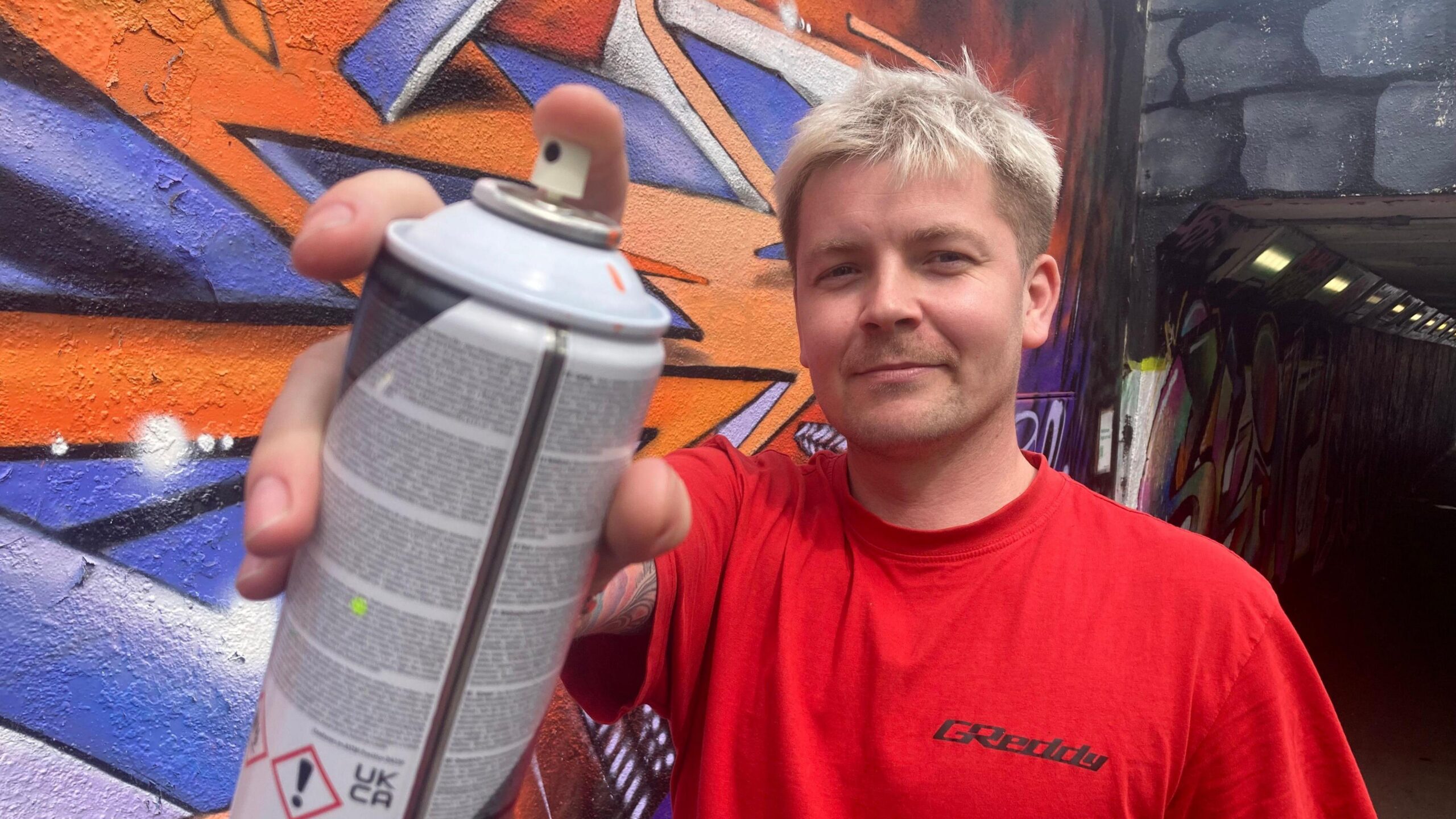 A man with dyed blonde hair wearing a red shirt holds a can of spray paint close to the camera as he stands in front of a piece of wall art.