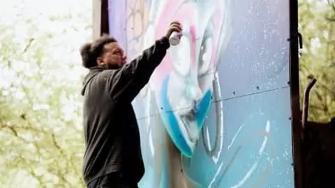 Handout A man in a black hoodie with afro hair in a bun is spraying paint onto a temporary wall. He is painting a woman's face in an animated but realistic style. There are trees behind him and the painting.